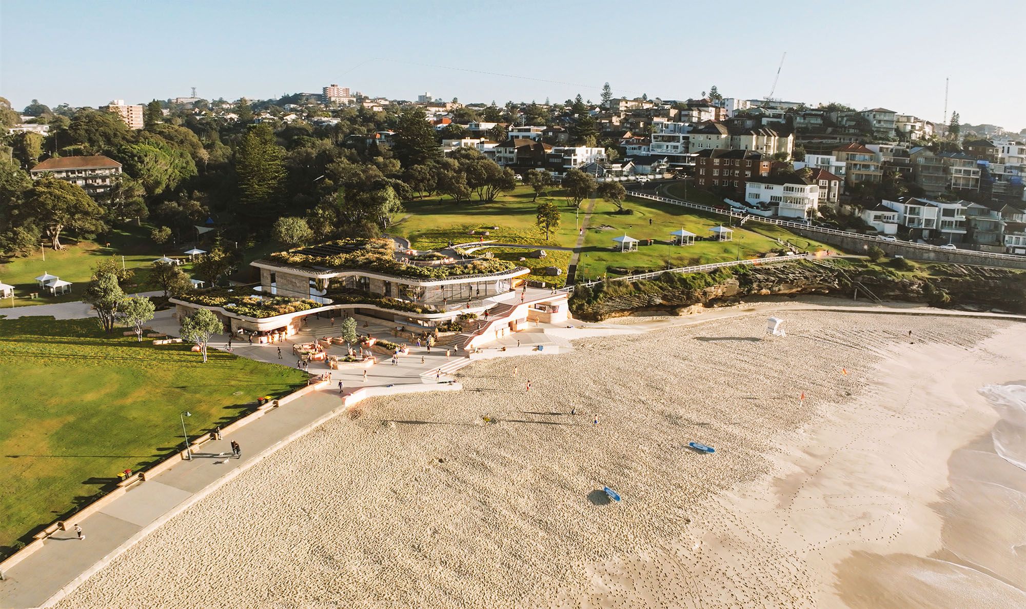A digital render of a beach, with a prominent contemporary, curved, glass building with green roof on the shore.