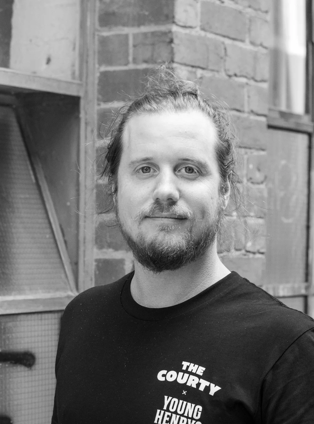 A black and white headshot of a person in-front of a brick wall, wearing a t-shirt that says 'The Courty'