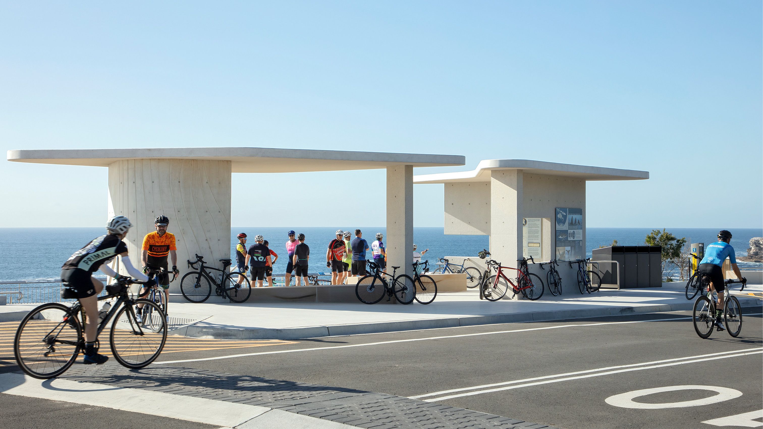 A photograph of a group of cyclist mingling around a lookout. Blue sea and Skies are behind. Two cyclists ride past on the road in front.  