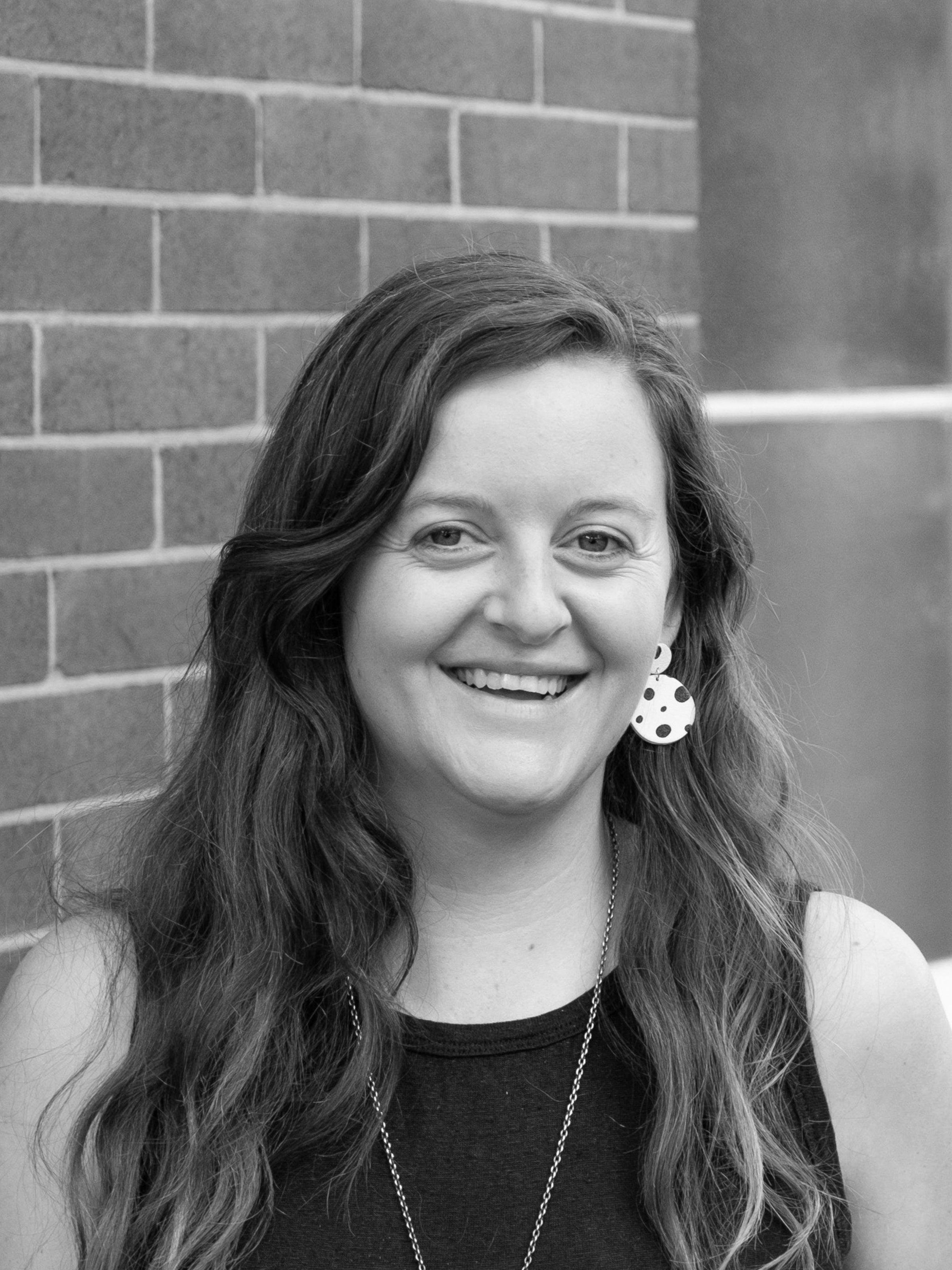 A black and white headshot of a smiling person wearing earrings, standing in front of a brick wall and window.