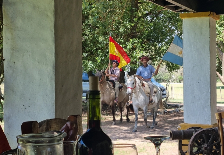 From a covered patio, two men on horseback carry Spanish and Argentinian flags past an old cannon.