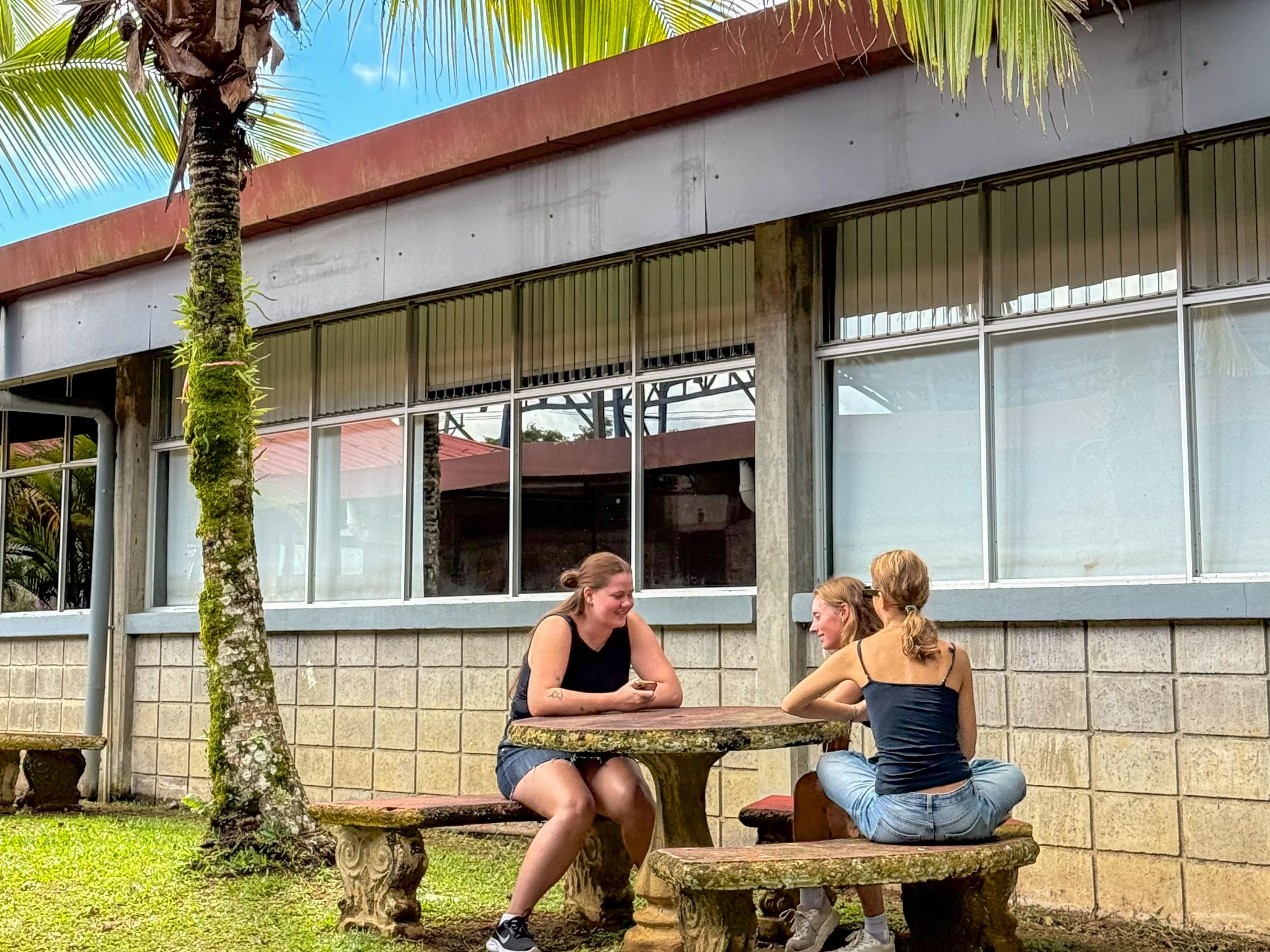Three women chatting at an outdoor stone table in front of a building.