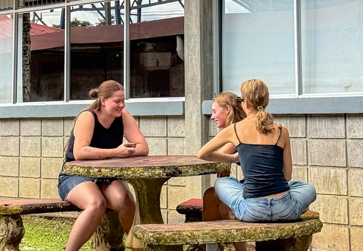 Three women chatting at an outdoor stone table in front of a building.