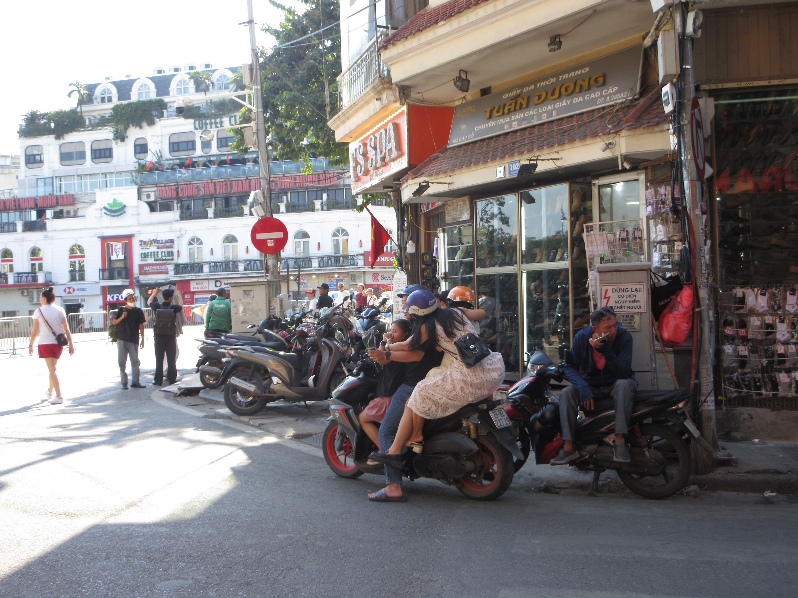 A sunny city street scene filled with parked motorcycles, people walking and sitting on bikes, and shops lining the road with multi-story buildings in the background.