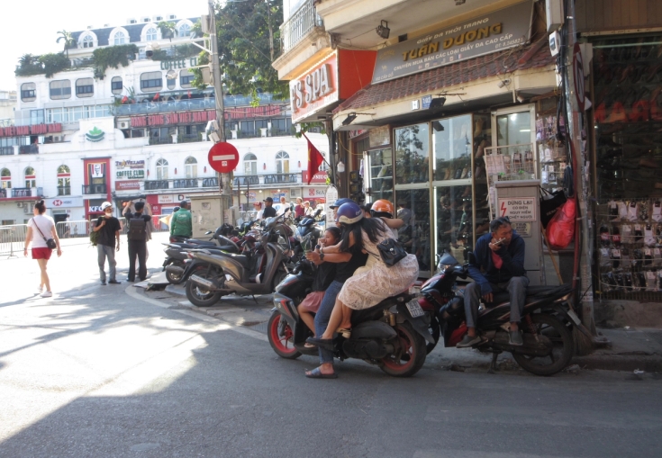 A sunny city street scene filled with parked motorcycles, people walking and sitting on bikes, and shops lining the road with multi-story buildings in the background.