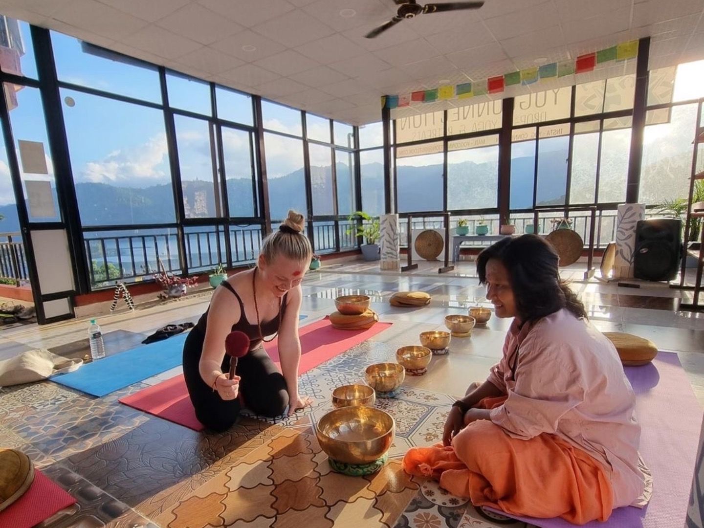 a woman is kneeling on a yoga mat next to a woman in a room with bowls on the floor.