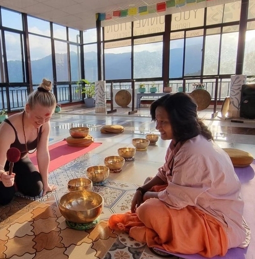 a woman is kneeling on a yoga mat next to a woman in a room with bowls on the floor.