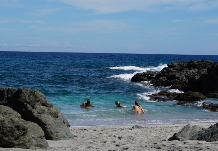 Three people swim in the clear turquoise water of a rocky beach on a sunny day.