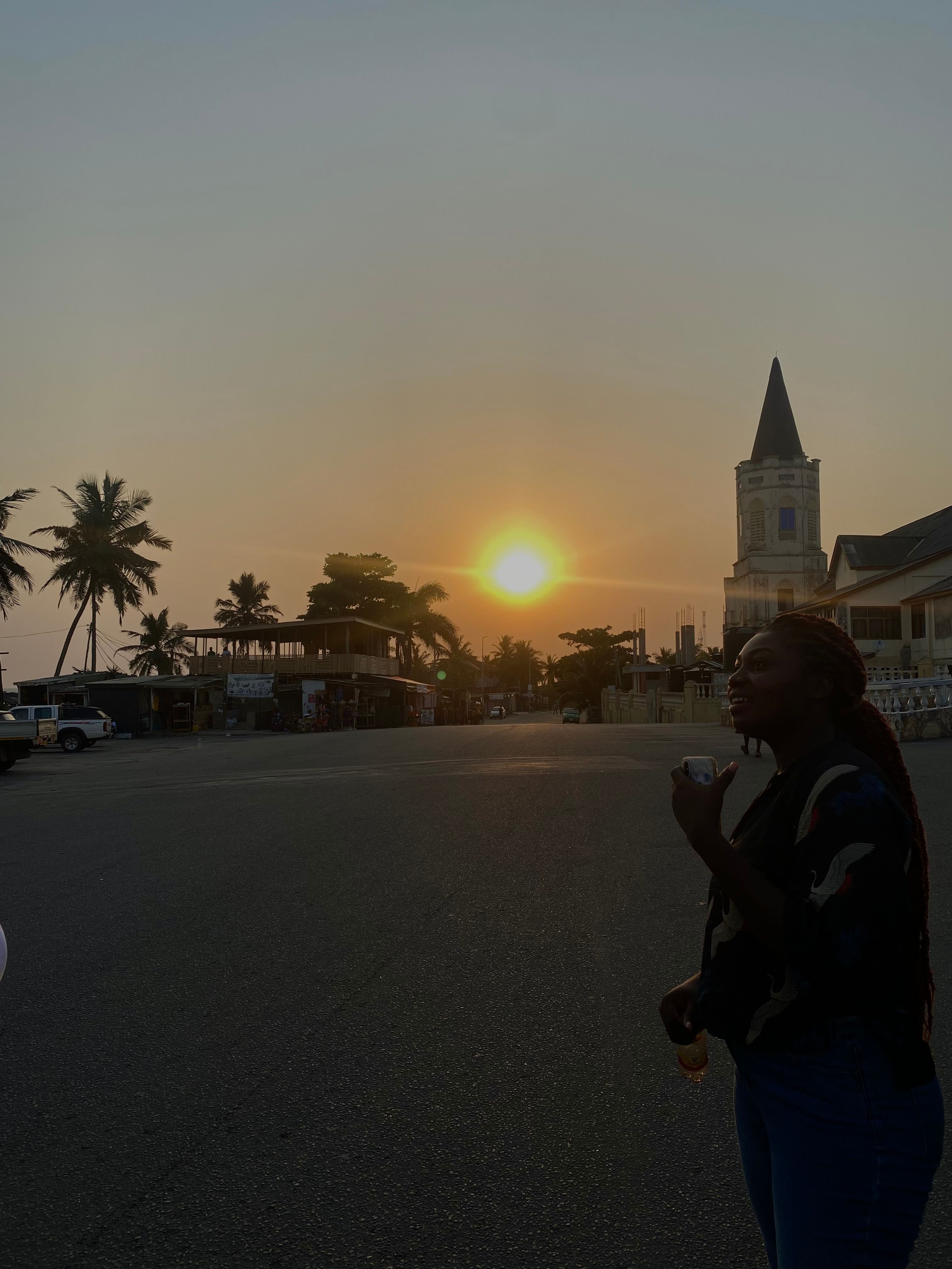 A silhouetted woman stands on a street at sunset, with palm trees and a tower in the background.