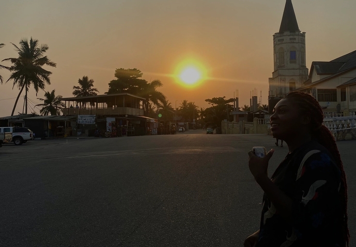 A silhouetted woman stands on a street at sunset, with palm trees and a tower in the background.