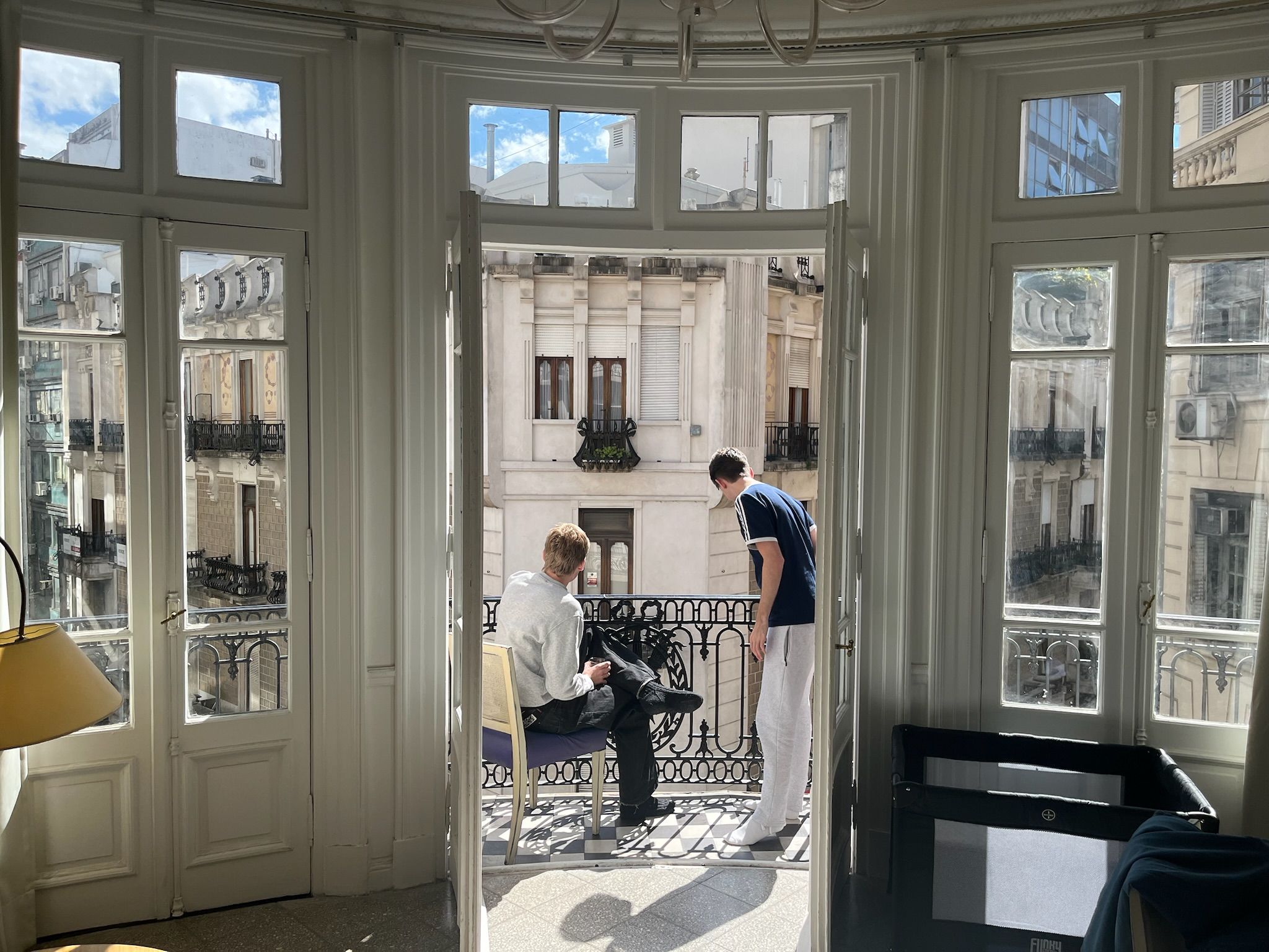 Two men on a sunny balcony overlooking a city street with ornate buildings.