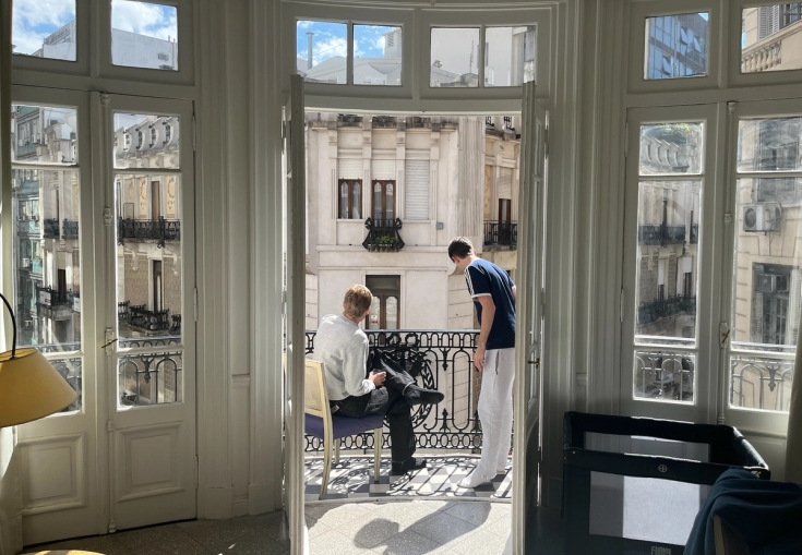 Two men on a sunny balcony overlooking a city street with ornate buildings.