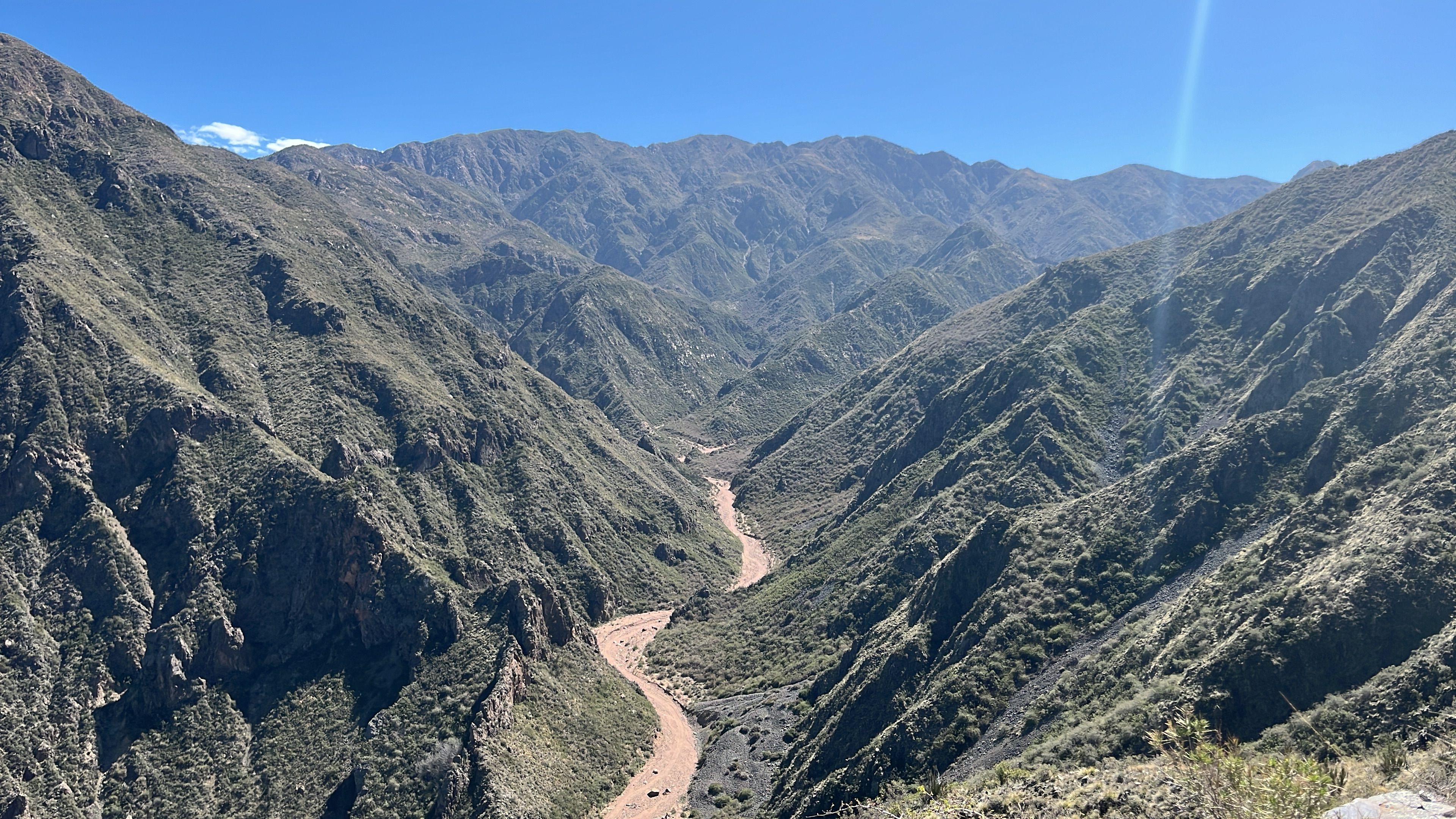 A reddish-brown river winds through a deep valley surrounded by steep, green mountains under a clear blue sky.