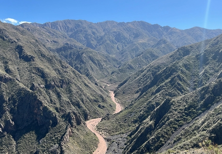 A reddish-brown river winds through a deep valley surrounded by steep, green mountains under a clear blue sky.