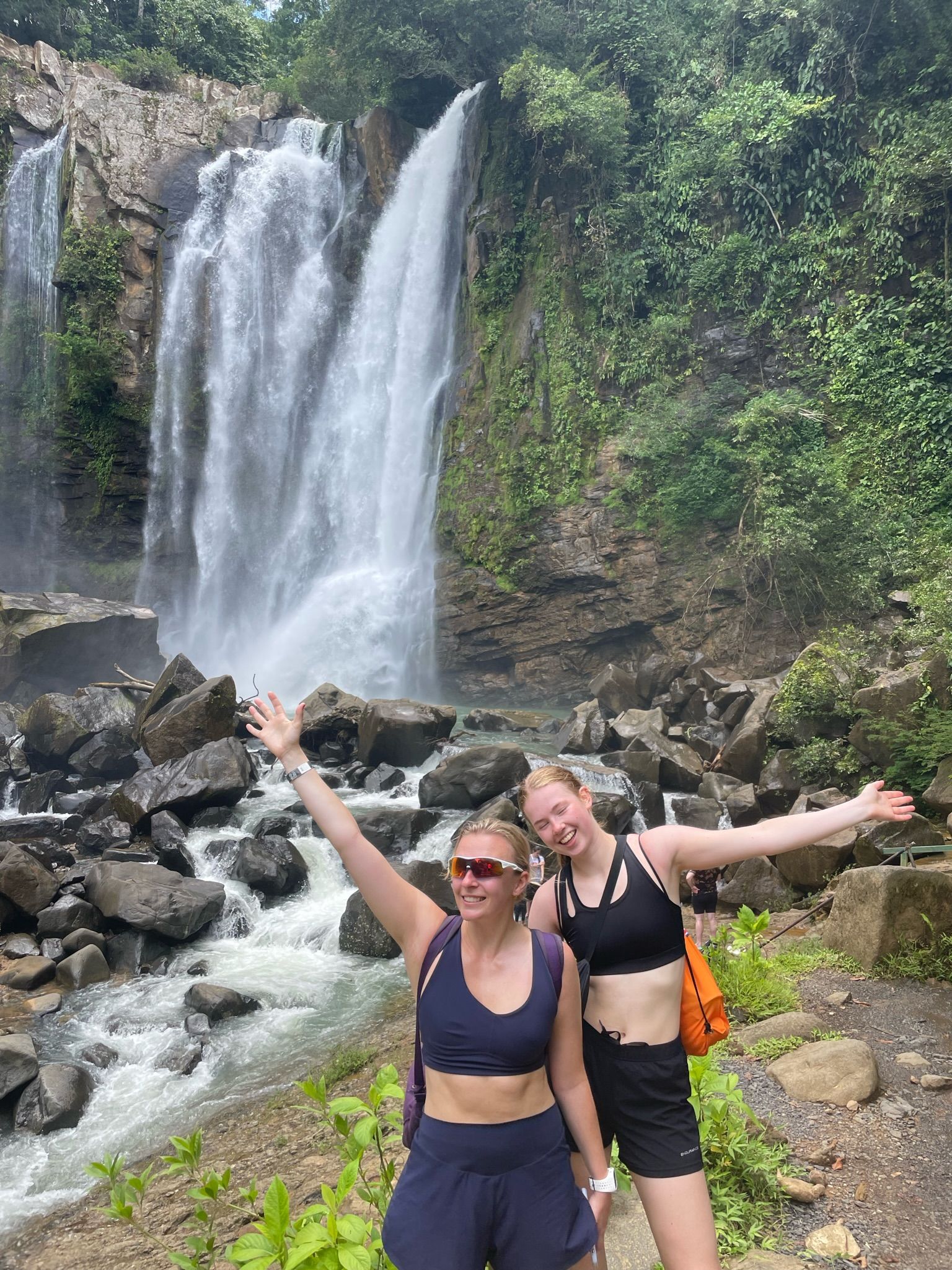 to kvinner står foran et fossefall med armene utstrakt.