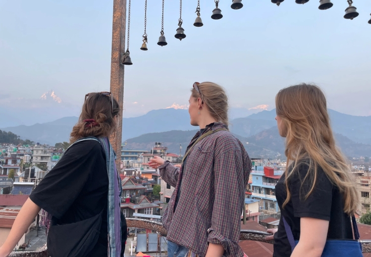Three women on a rooftop look at a city and snow-capped mountains, with bells hanging overhead.