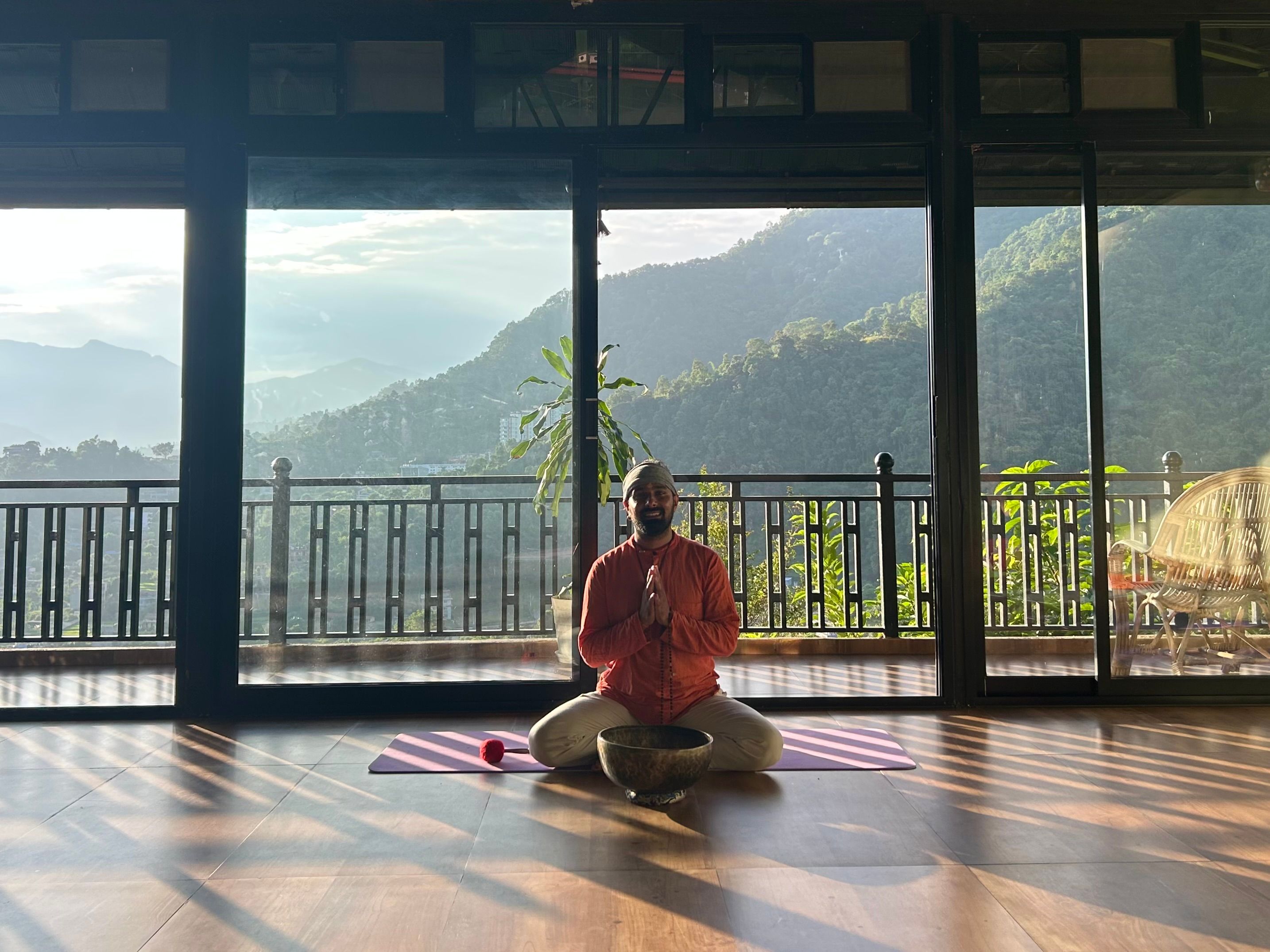 A man meditates cross-legged on a yoga mat with a singing bowl, in a sunlit room overlooking green mountains.