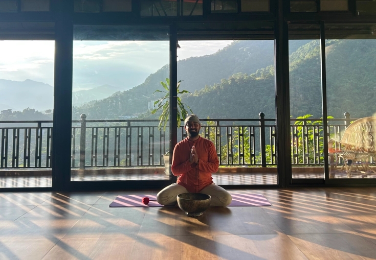 A man meditates cross-legged on a yoga mat with a singing bowl, in a sunlit room overlooking green mountains.