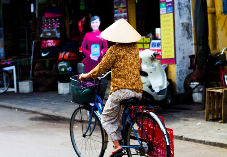 en kvinne som bærer en konisk hatt, sykler ned en gate.