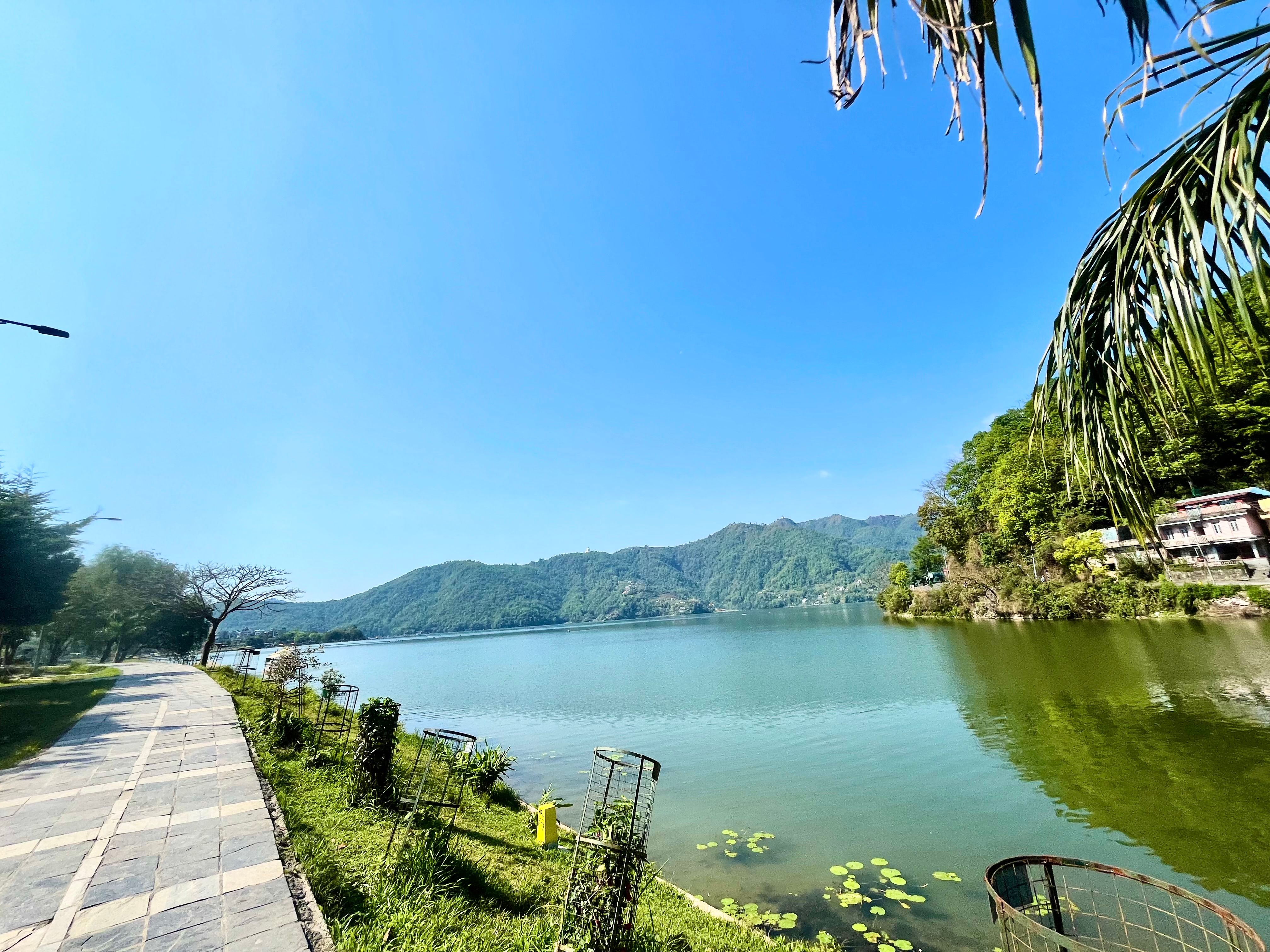 A paved path runs along a lake with distant mountains and houses under a clear blue sky.