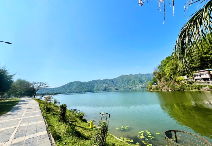 A paved path runs along a lake with distant mountains and houses under a clear blue sky.