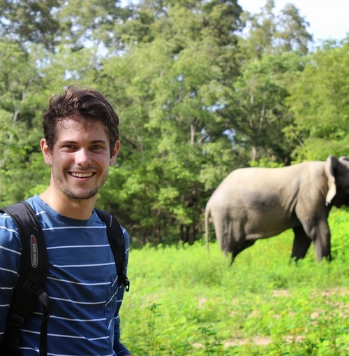 en man med en ryggsäck står på ett fält med en elefant i bakgrunden