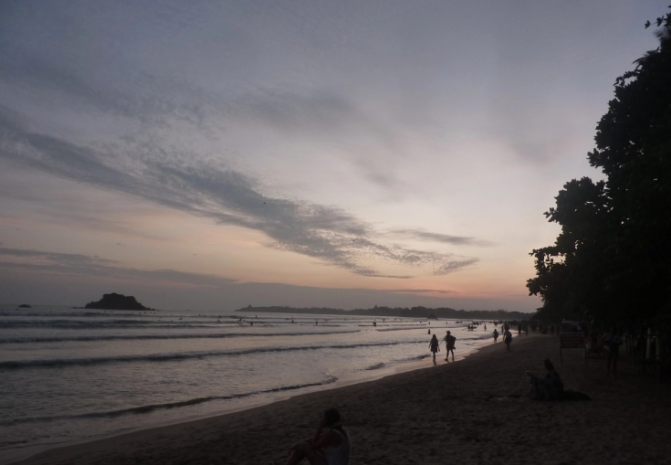 Beach at dusk with people walking and swimming, an island in the distance, under a cloudy, pastel sky.