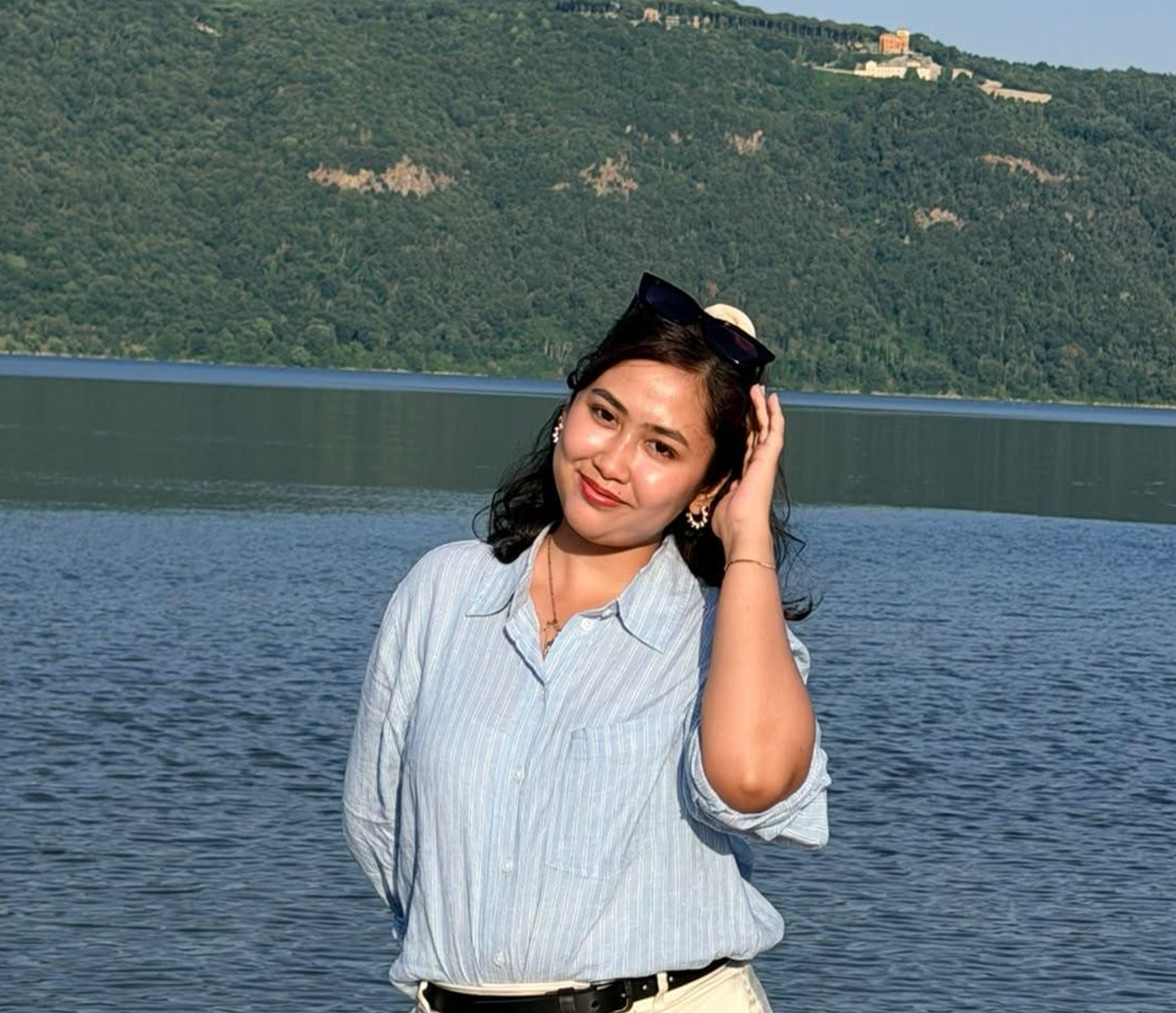 A smiling woman in a striped shirt stands by a lake with a forested mountain in the background.