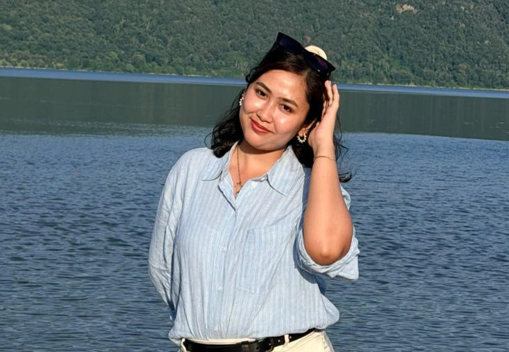 A smiling woman in a striped shirt stands by a lake with a forested mountain in the background.