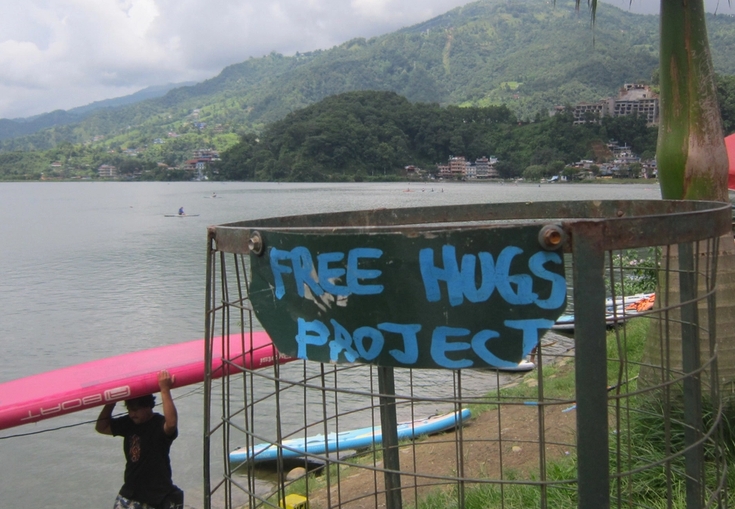 A "FREE HUGS PROJECT" sign by a lake with green mountains, as a man carries a pink paddleboard.