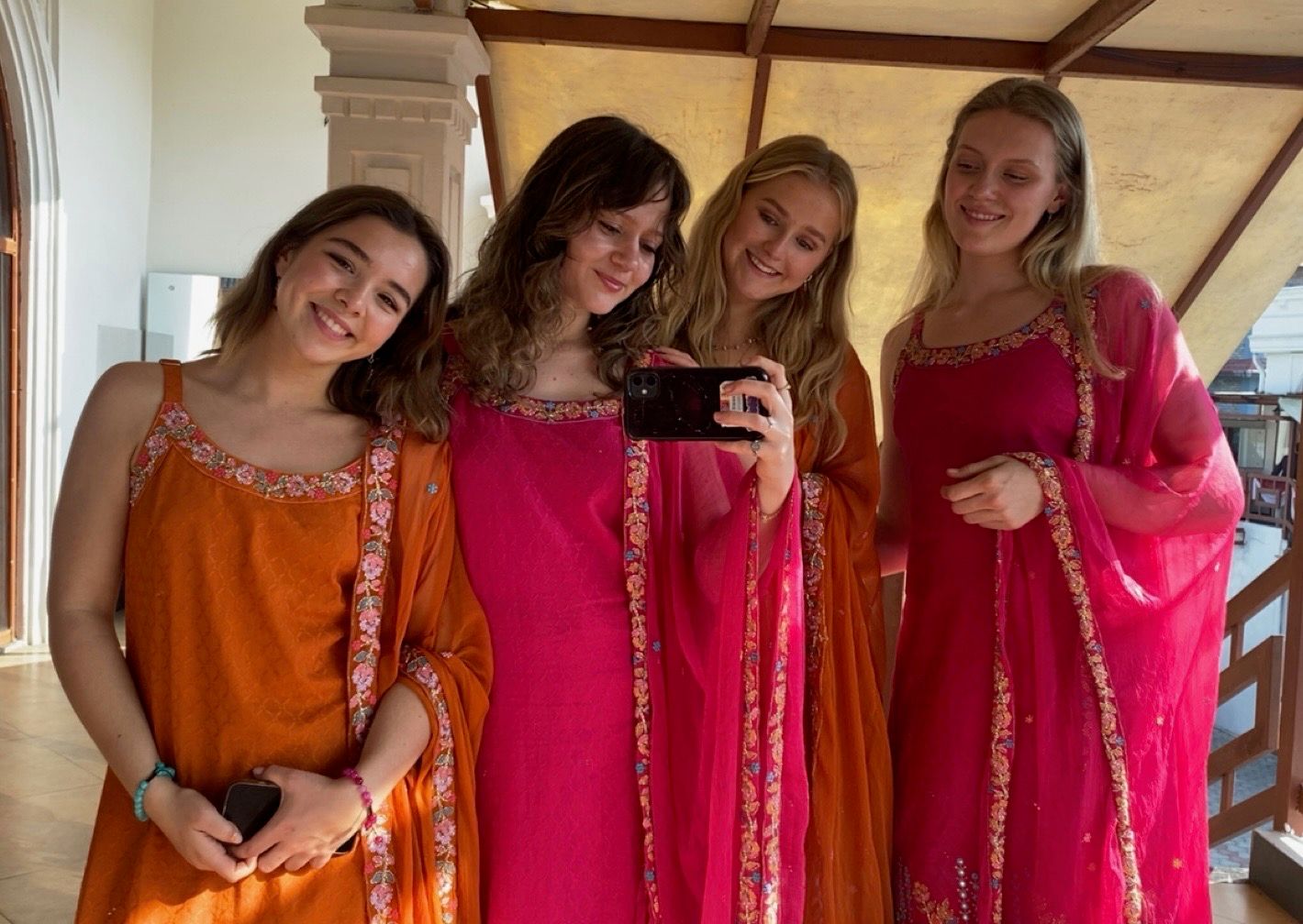 Four smiling young women in vibrant pink and orange traditional Indian attire taking a selfie.
