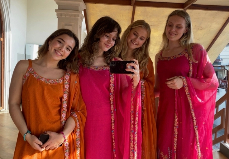 Four smiling young women in vibrant pink and orange traditional Indian attire taking a selfie.