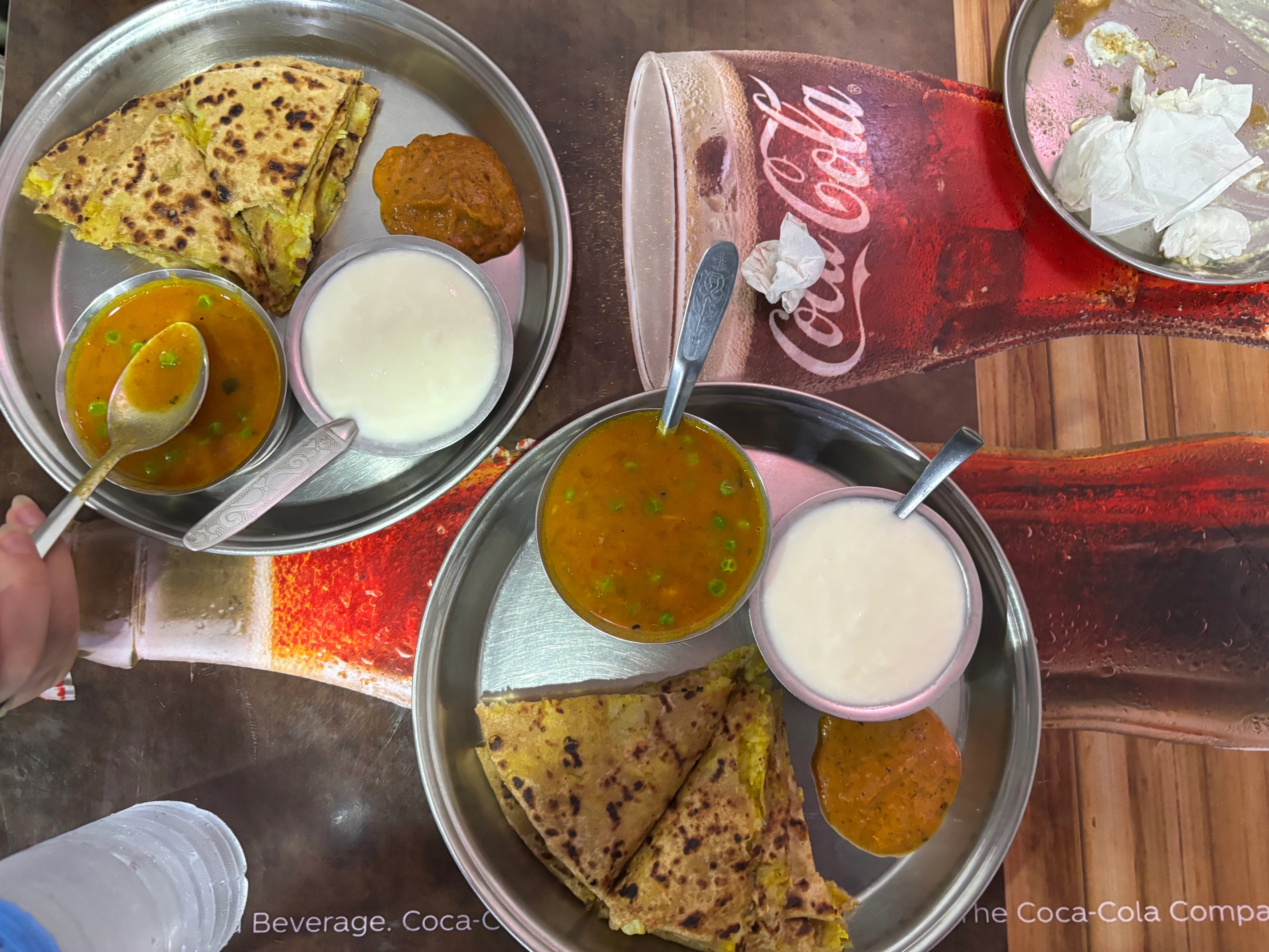 Two metal thalis with Indian food (paratha, curry, yogurt) on a Coca-Cola tablecloth, one partially eaten.