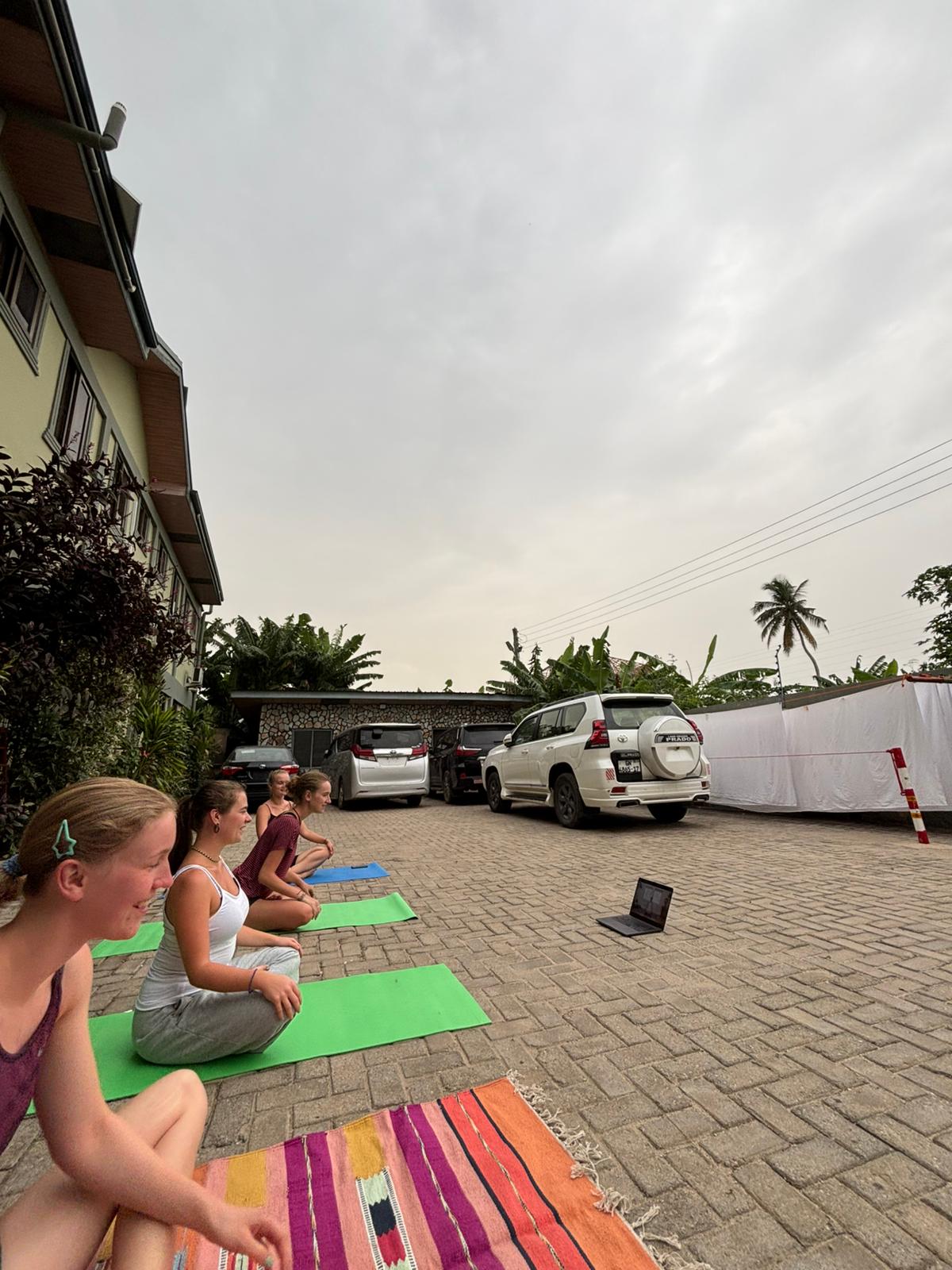 Four young women sit on yoga mats and a colorful rug outdoors, watching a laptop with parked cars behind them.