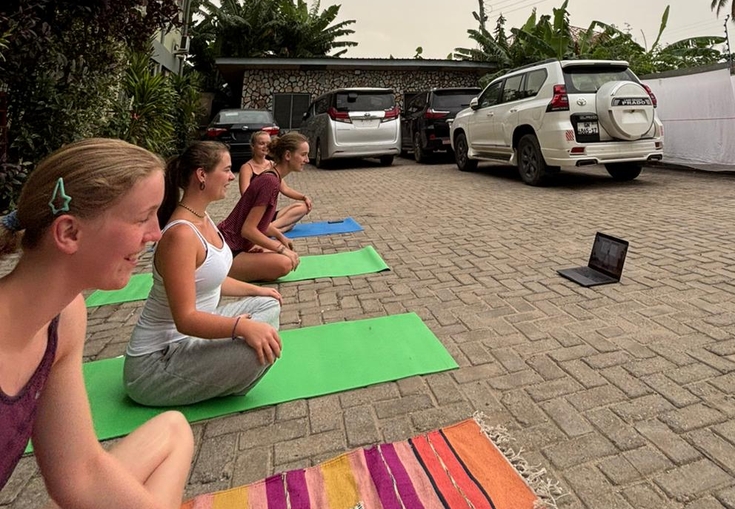 Four young women sit on yoga mats and a colorful rug outdoors, watching a laptop with parked cars behind them.