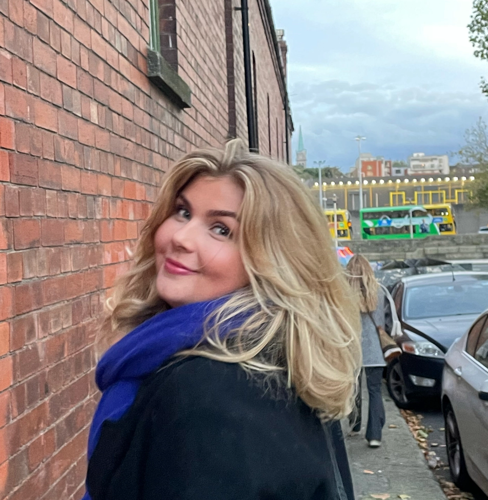 a woman wearing a black coat and a blue scarf stands in front of a brick wall