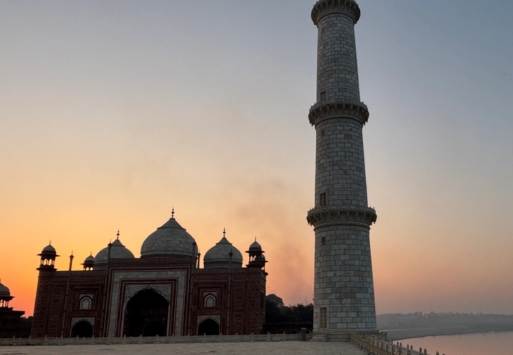 A red sandstone building and a tall white minaret stand against a colorful sunrise sky, with a river in the distance.