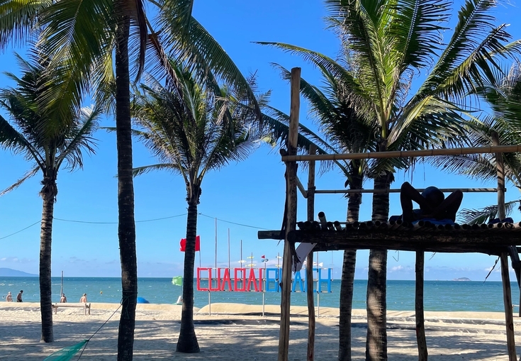 A person relaxes on a wooden platform on a sunny beach with palm trees and a colorful "CUA DAI BEACH" sign in the distance.