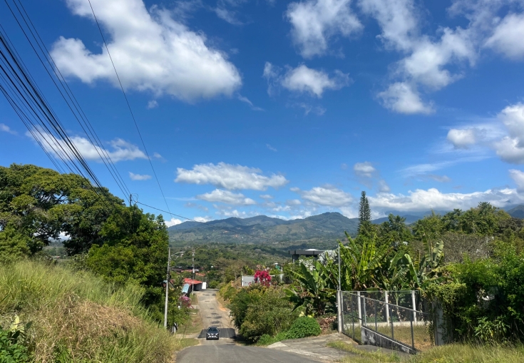 A paved road slopes down through a green landscape with mountains in the distance under a blue sky with white clouds.