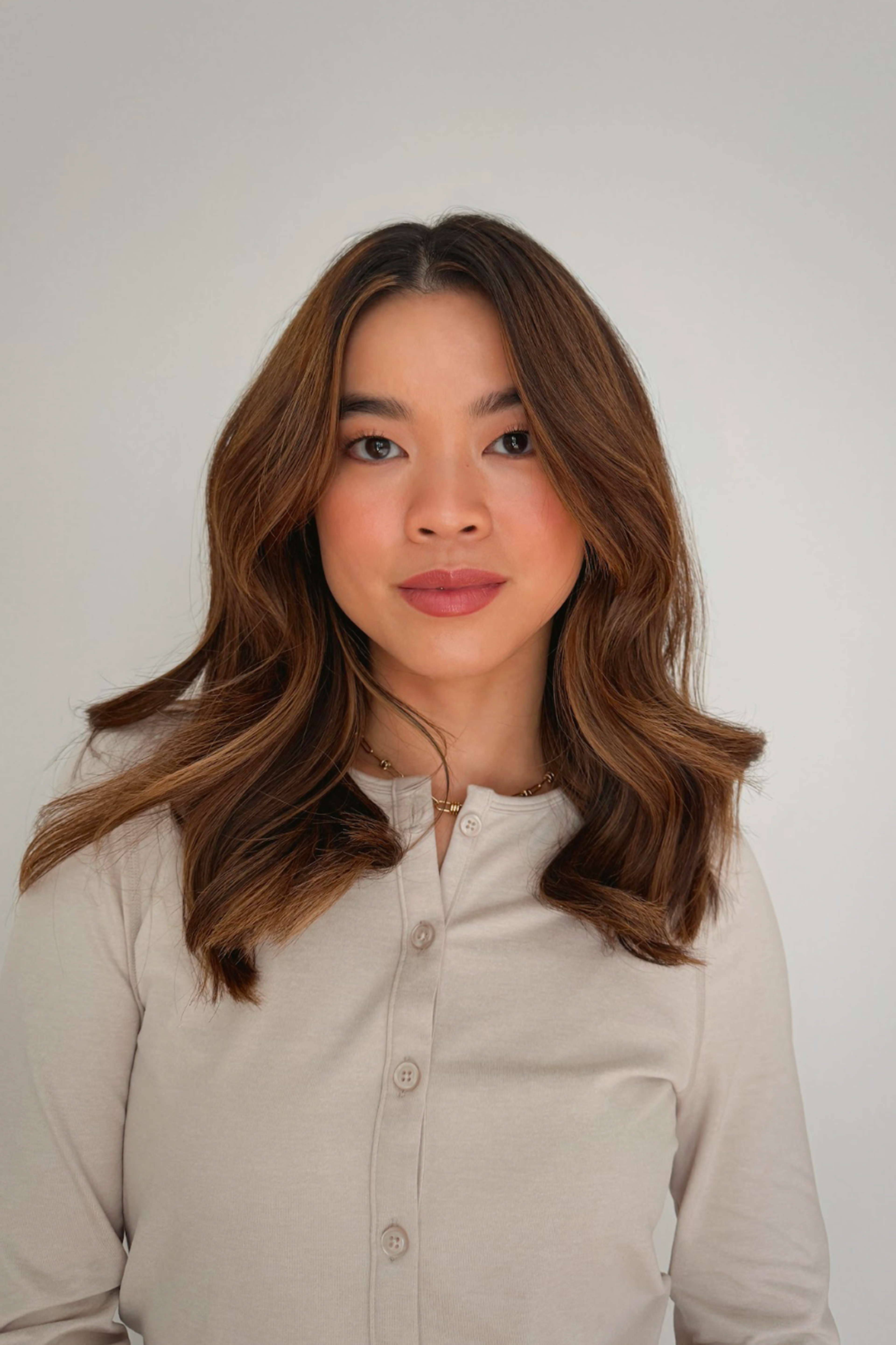 A woman with wavy brown hair wearing a beige button-up top stands against a plain white background, looking directly at the camera with a neutral expression.