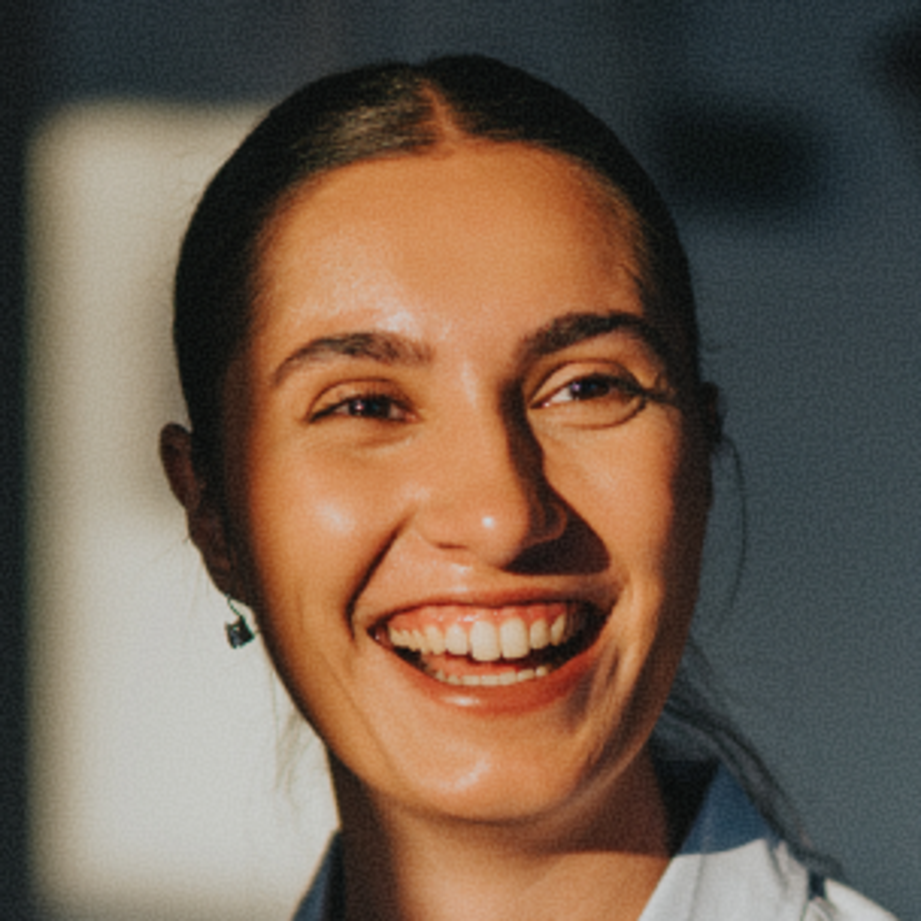 A woman with dark hair pulled back smiles brightly in warm sunlight, wearing a light-colored top and small earrings.