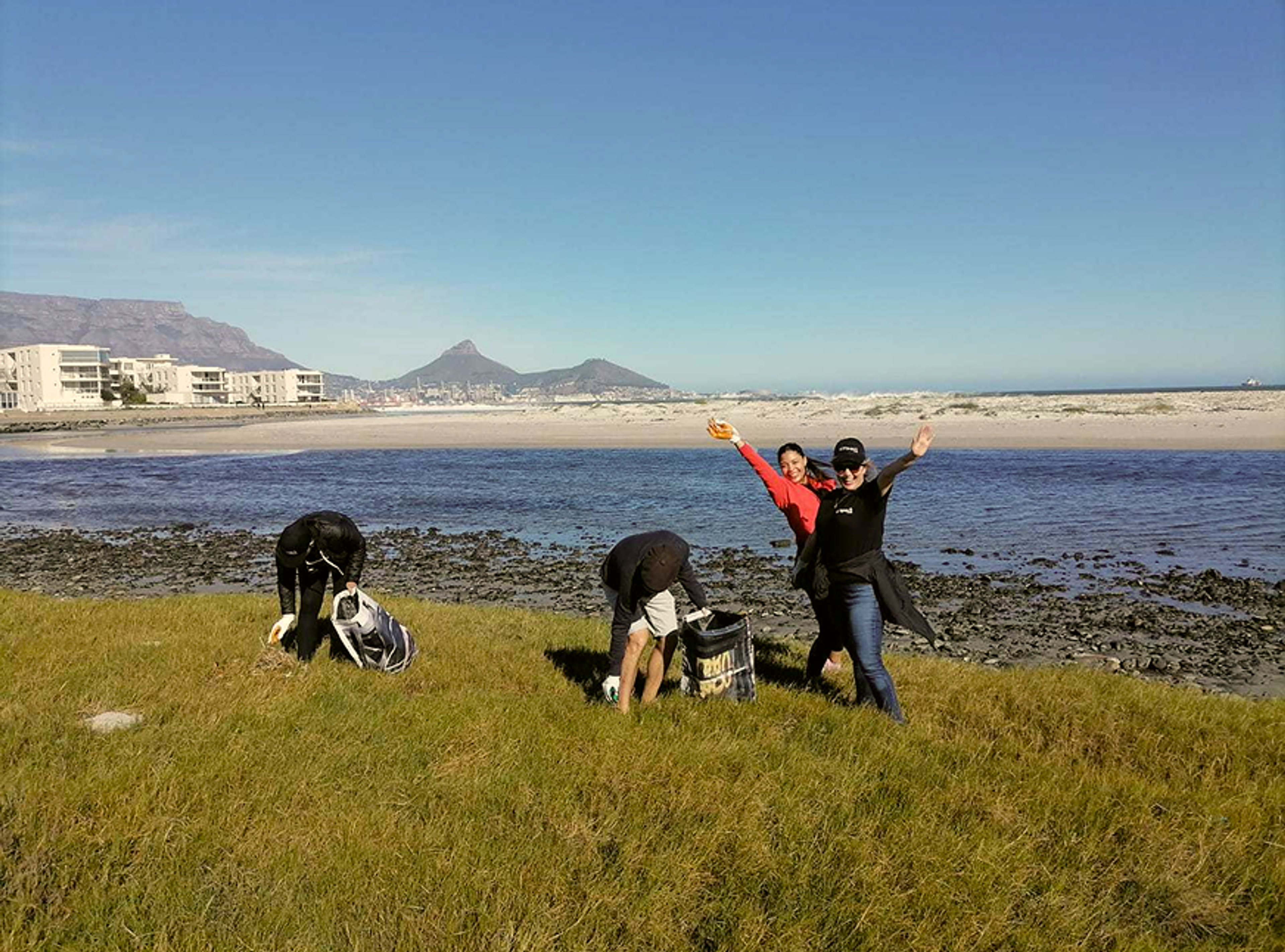 Four people clean up litter on a grassy shore near the beach. Two are bent over picking up trash, while two others stand, smiling and posing with arms raised. Ocean, buildings, and mountains are visible in the background.