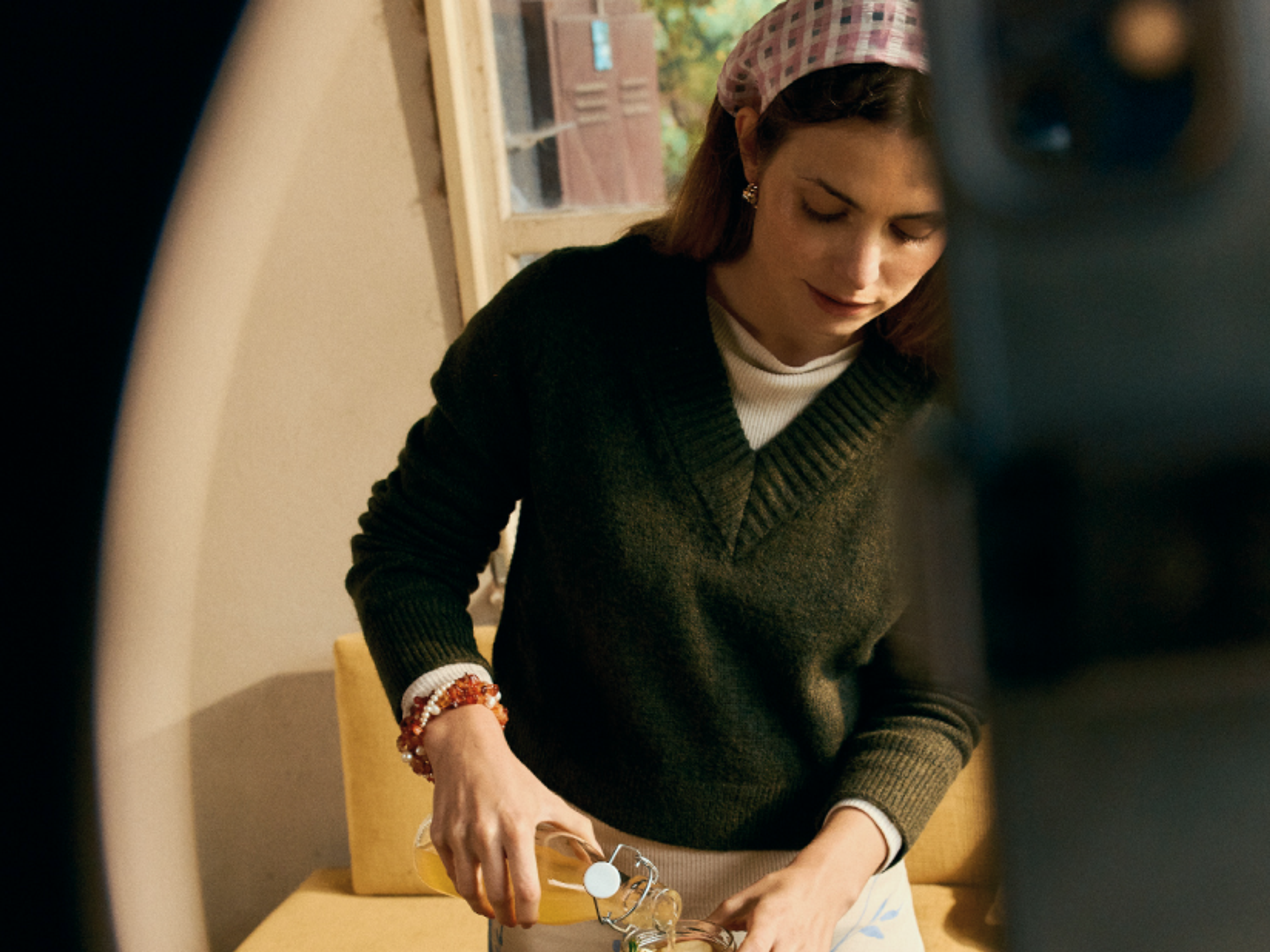 A woman wearing a dark green sweater and a pink headband pours a drink into a glass at a table indoors, with soft light coming from a nearby window.