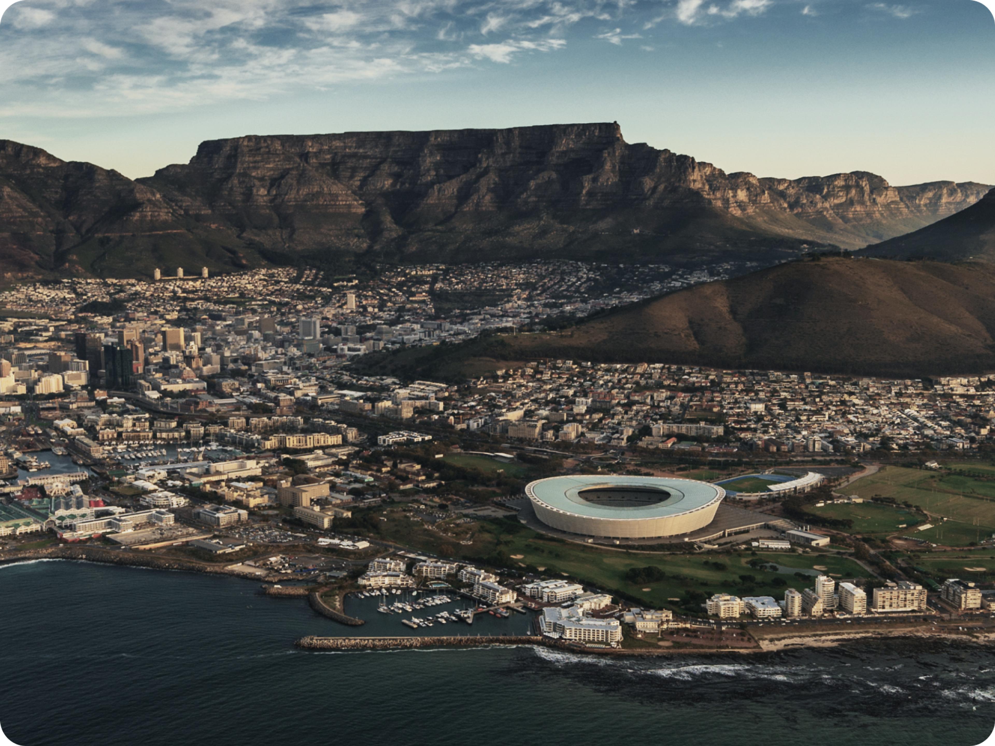 Aerial view of Cape Town, South Africa, showing the city, Cape Town Stadium, the coastline, and Table Mountain in the background under a partly cloudy sky.