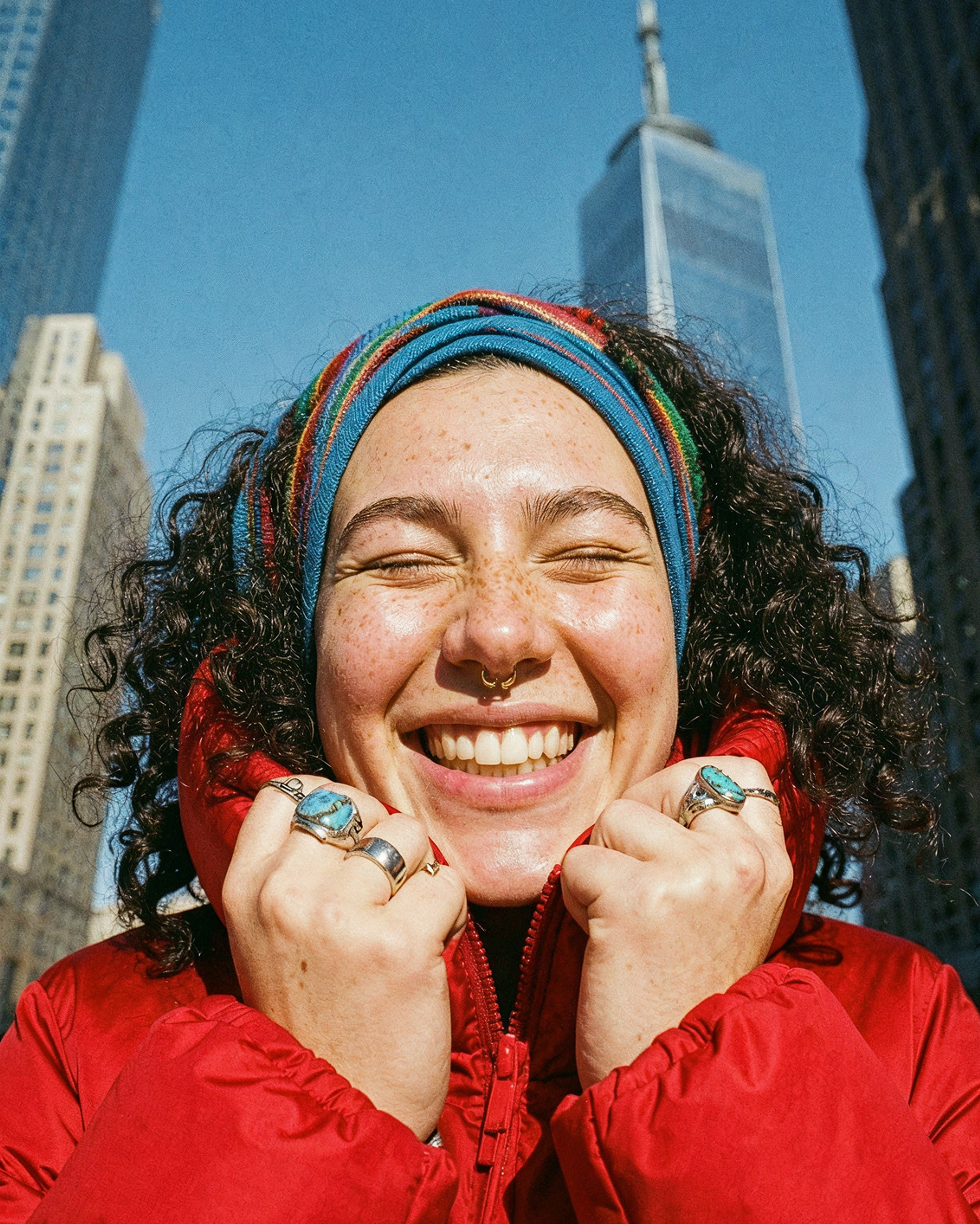 A smiling person with curly hair, colorful headband, rings, and red jacket stands outdoors in a city with tall buildings under a clear blue sky.