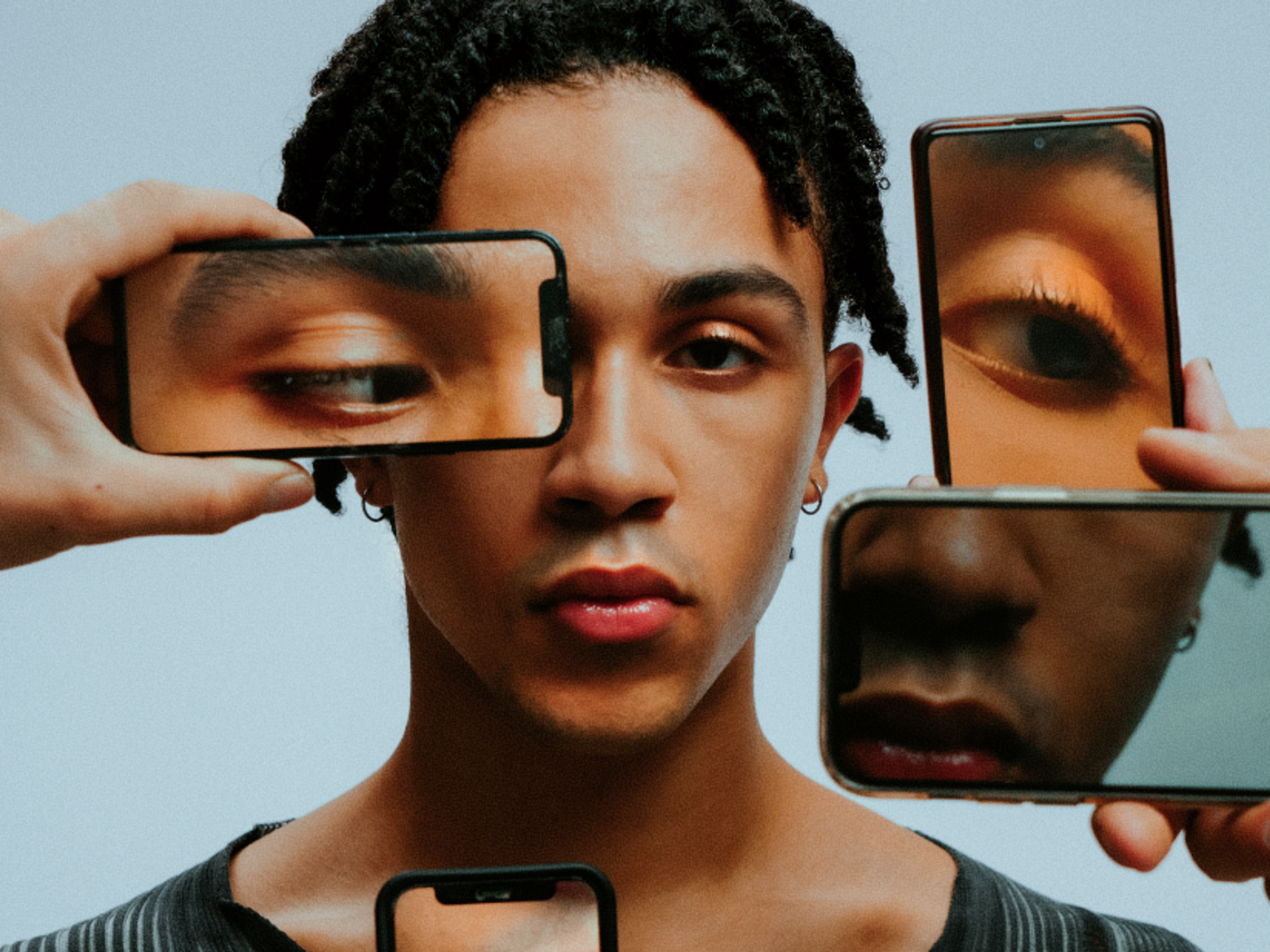 A person with medium skin tone is surrounded by four smartphones showing close-up images of their eyes and mouth, partially covering their face against a plain background.
