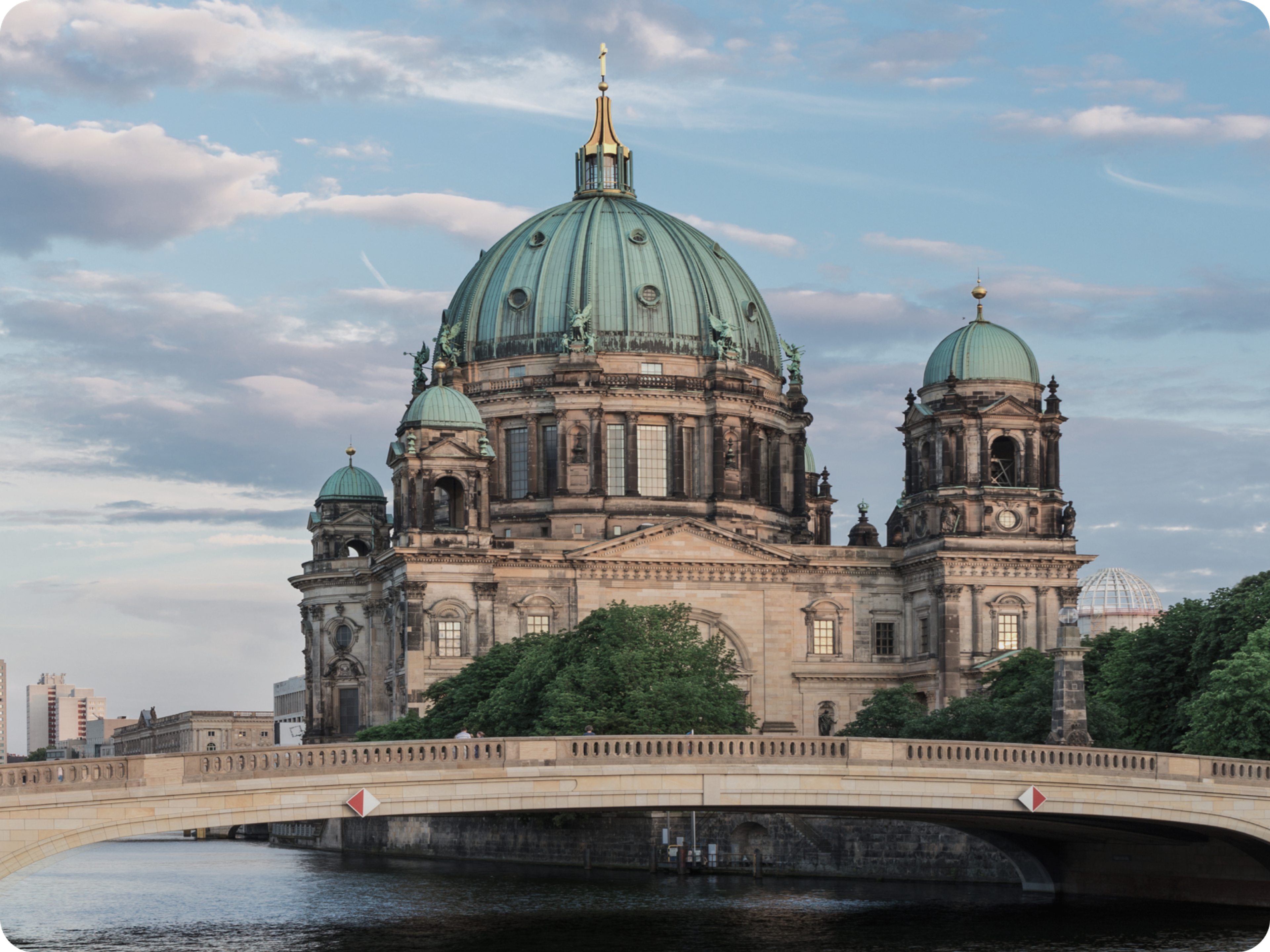 Berlin Cathedral with its large green dome and two smaller towers, seen at sunset behind a stone bridge over the river, with trees in the foreground and a partly cloudy sky above.
