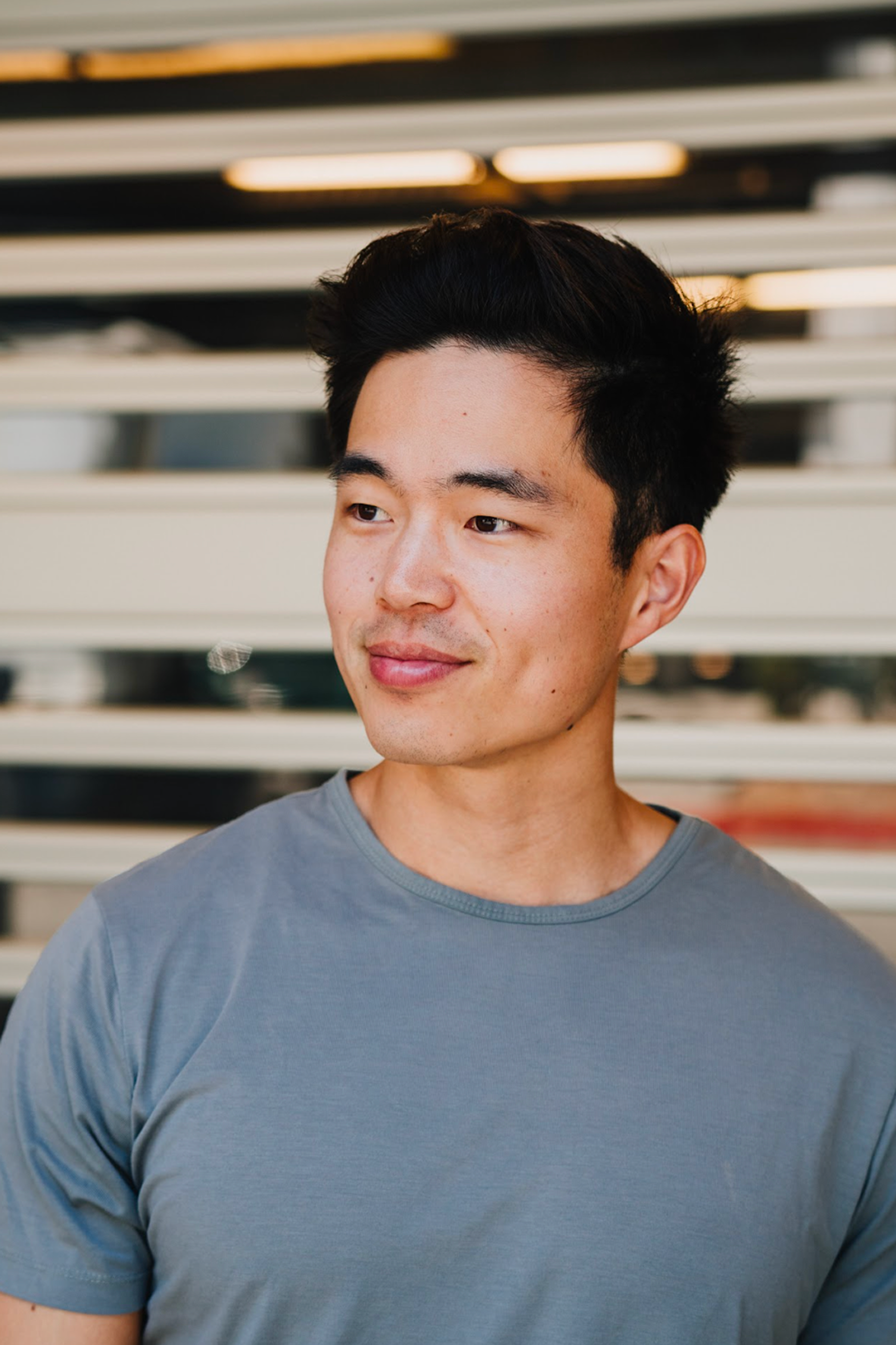 A young man with short black hair wearing a gray t-shirt stands in front of a striped background, looking slightly to the side and smiling softly.