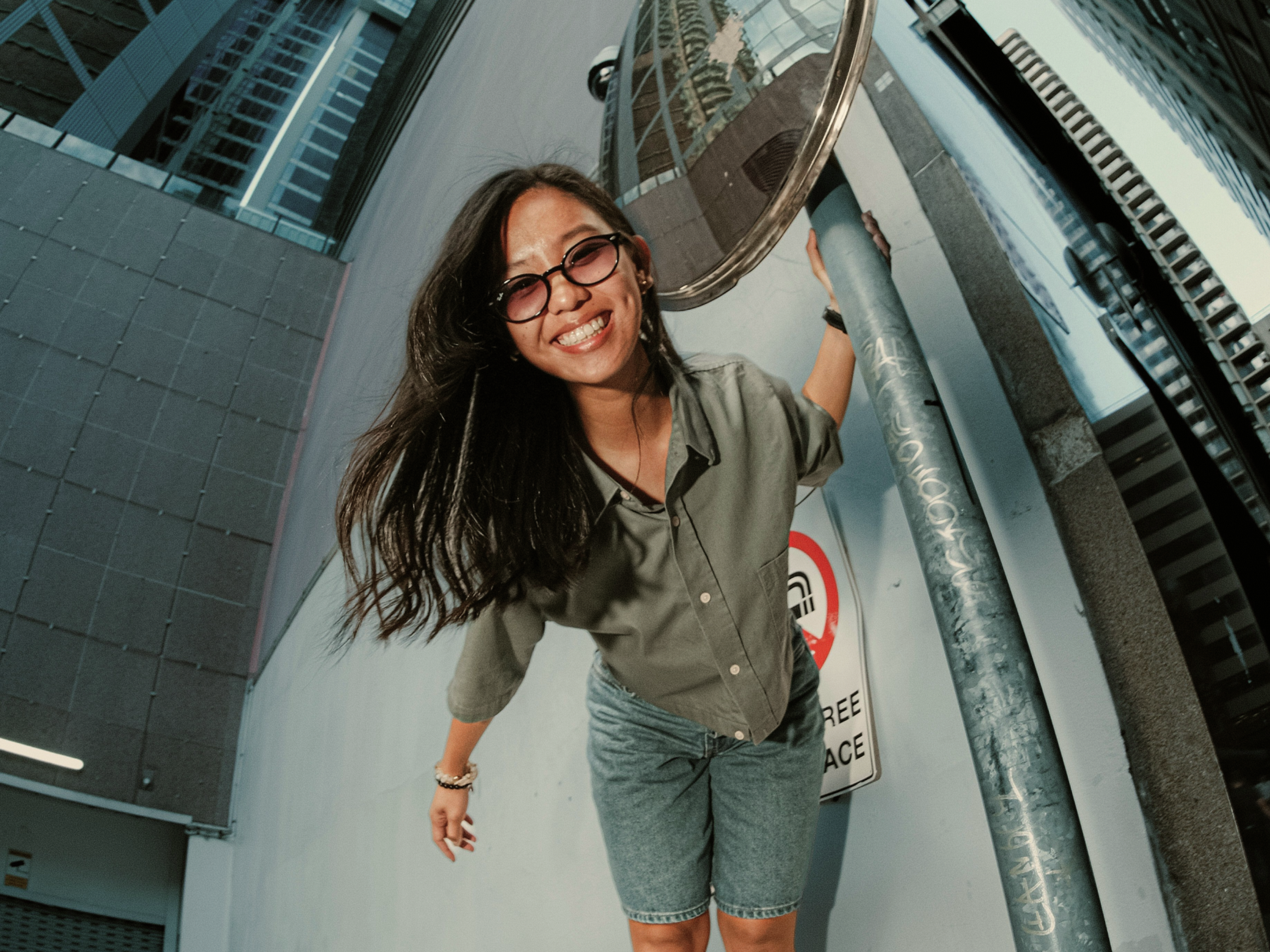 A smiling woman with glasses stands outdoors, leaning on a metal pole beside a convex mirror. She wears a green shirt and denim shorts, with buildings reflected in the mirror behind her.