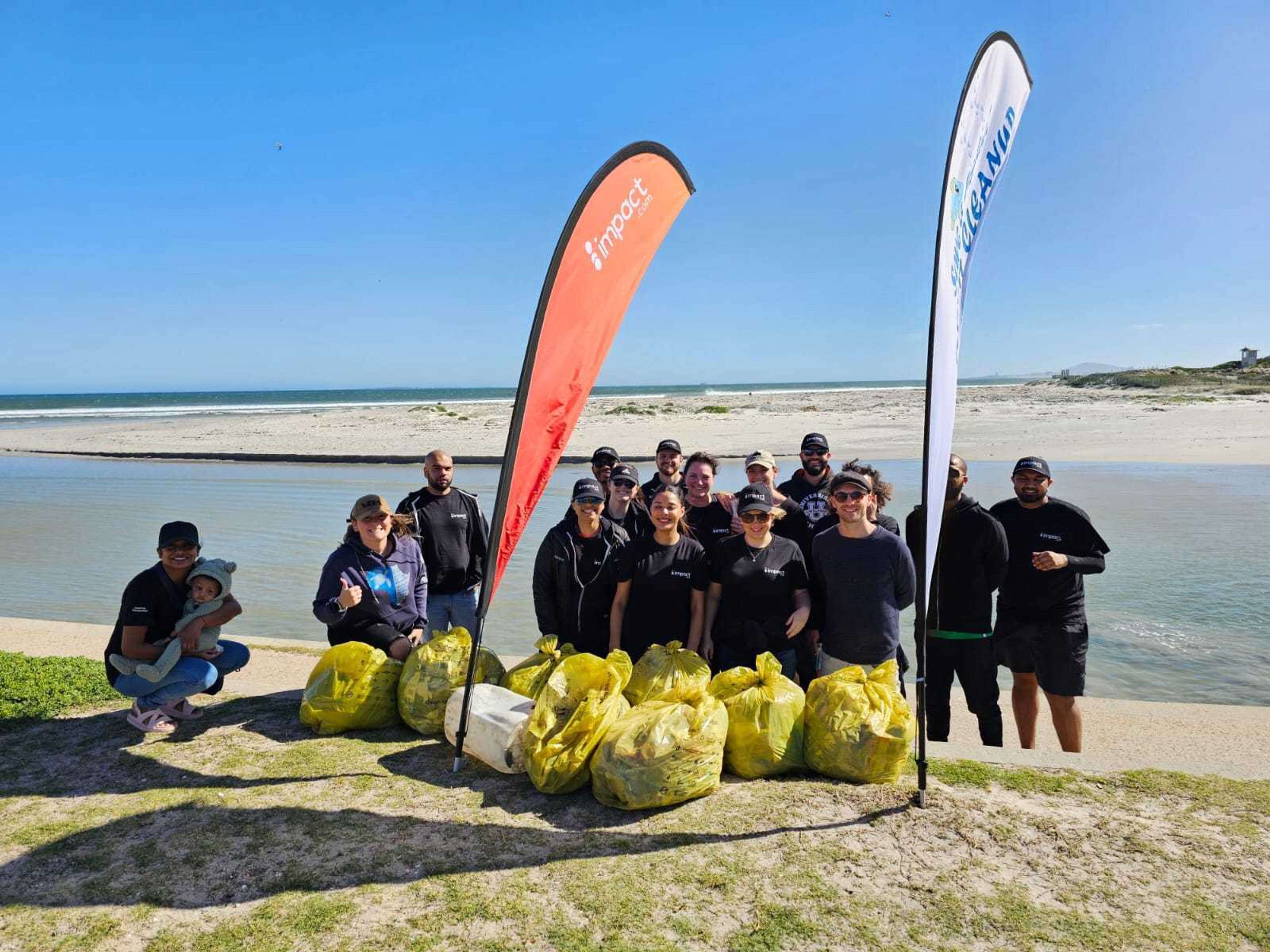 A group of people poses on a beach behind bags of collected trash, with two tall flags and water in the background. The scene is sunny and everyone is smiling.